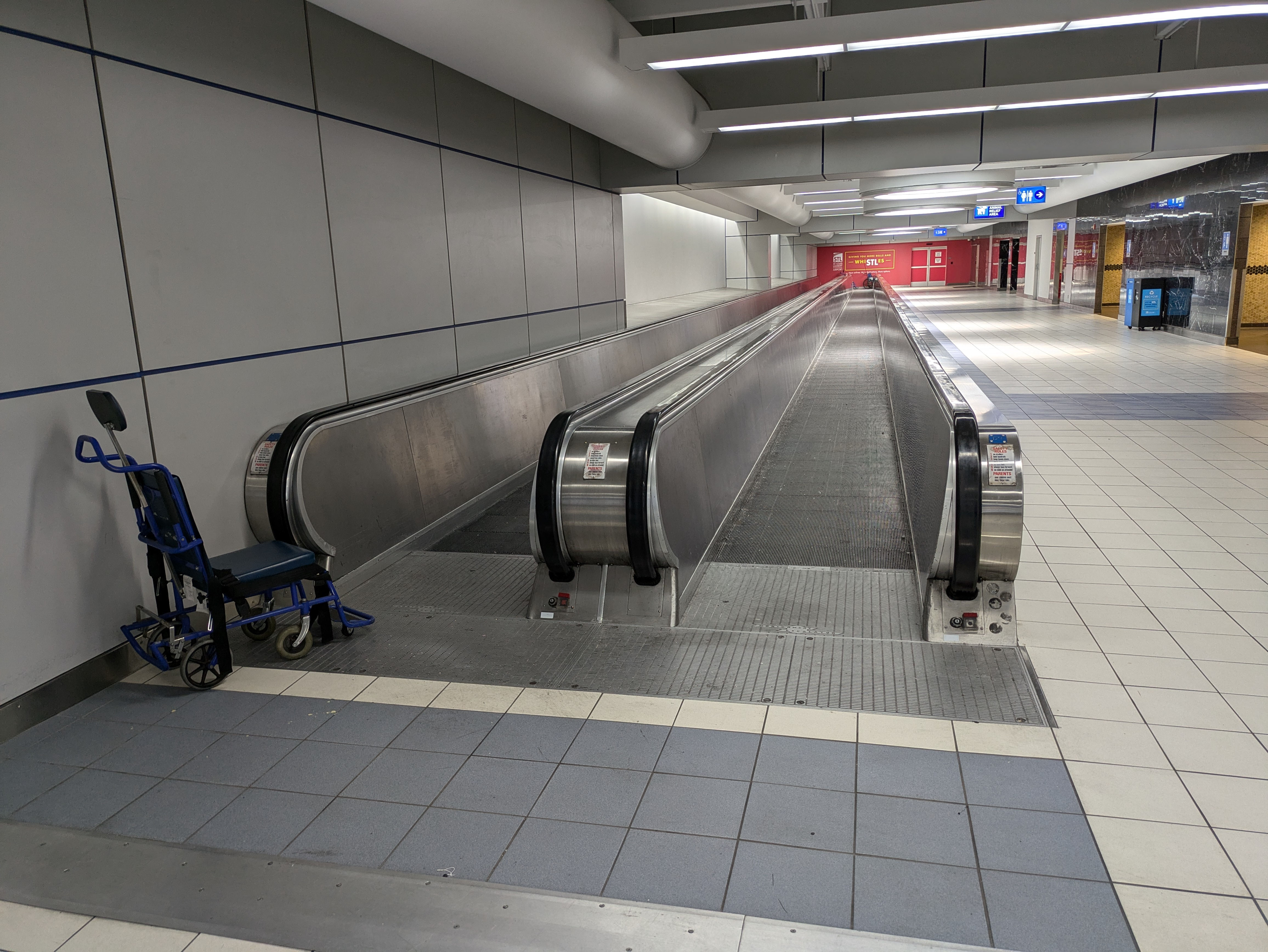 A moving sidewalk leading to a red wall, with "Giving you more bells and whiSTLes" optimistically painted on. The bathrooms can be seen to the right, with nobody entering or leaving. The whole area is eerily vacant.